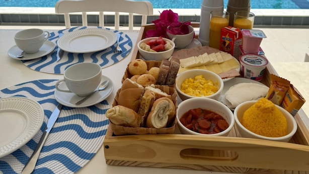 A breakfast tray with various foods and drinks on a table by a pool with a view of the ocean and resort buildings. - KEFI POUSADA (Maragogi)