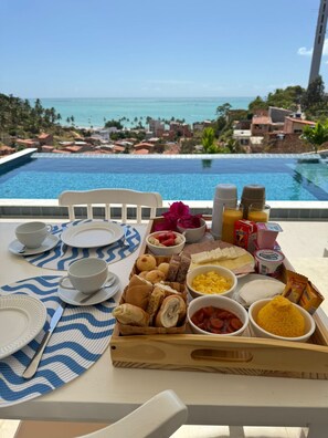 A breakfast tray with various foods and drinks on a table by a pool with a view of the ocean and resort buildings. - KEFI POUSADA (Maragogi)