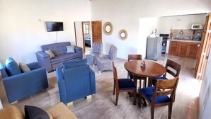 Ein geräumiger Wohnbereich mit blauem Sofa, einem Holzesstisch mit Stühlen, einer Küchenzeile inklusive Mikrowelle und einem an der Wand befestigten Fernseher.