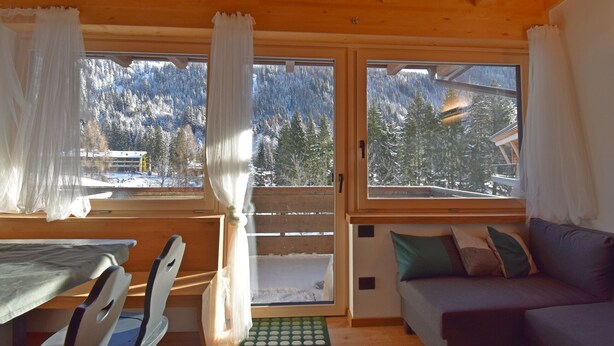 Ein gemütliches Blockhaus-Interieur mit Holzbalkendecke, einem Esstisch mit Stühlen, einer Couch mit Kissen und großen Fenstern mit Blick auf schneebedeckte Berge.