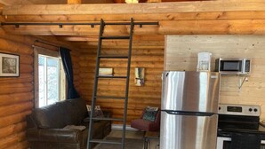 A log cabin interior with a wooden ceiling, a black metal ladder, a stainless steel refrigerator, a microwave, and a stove.