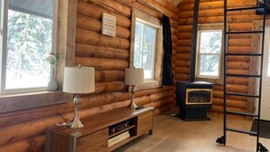 A log cabin interior with a wooden wall, a fireplace, a ladder, and a window with a view of trees.
