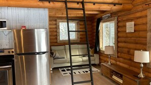 A log cabin interior with a stainless steel refrigerator, a wooden ladder, a sofa, and a wooden cabinet with a lamp.
