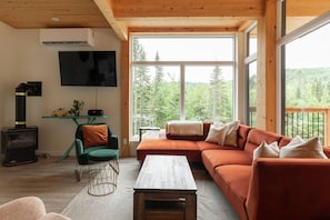 A cozy living room with a sofa, a television, a fireplace, and a large window overlooking a forest.
