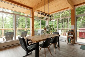 A spacious room with a wooden table, chairs, and large windows offering a forest view.
