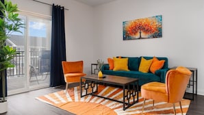 Ein modernes Wohnzimmer mit einem türkisfarbenen Sofa, orangefarbenen Sesseln, einem Holztisch und einem großen Fenster mit Blick auf einen Balkon.
