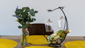 Une table à manger avec des sets de table jaunes, un vase contenant des feuilles vertes et une bouteille de vin posée sur un porte-bouteilles en fil de fer.