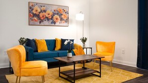 Un salon avec un canapé bleu, des fauteuils jaunes, une table basse en bois et un tableau représentant des fleurs accroché au mur.