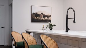 Moderni keittiö, jossa on valkoinen työtaso, kolme vihreillä tyynyillä varustettua punottua baarituolia, musta hana ja seinällä oleva kehystetty hevosten kuva.