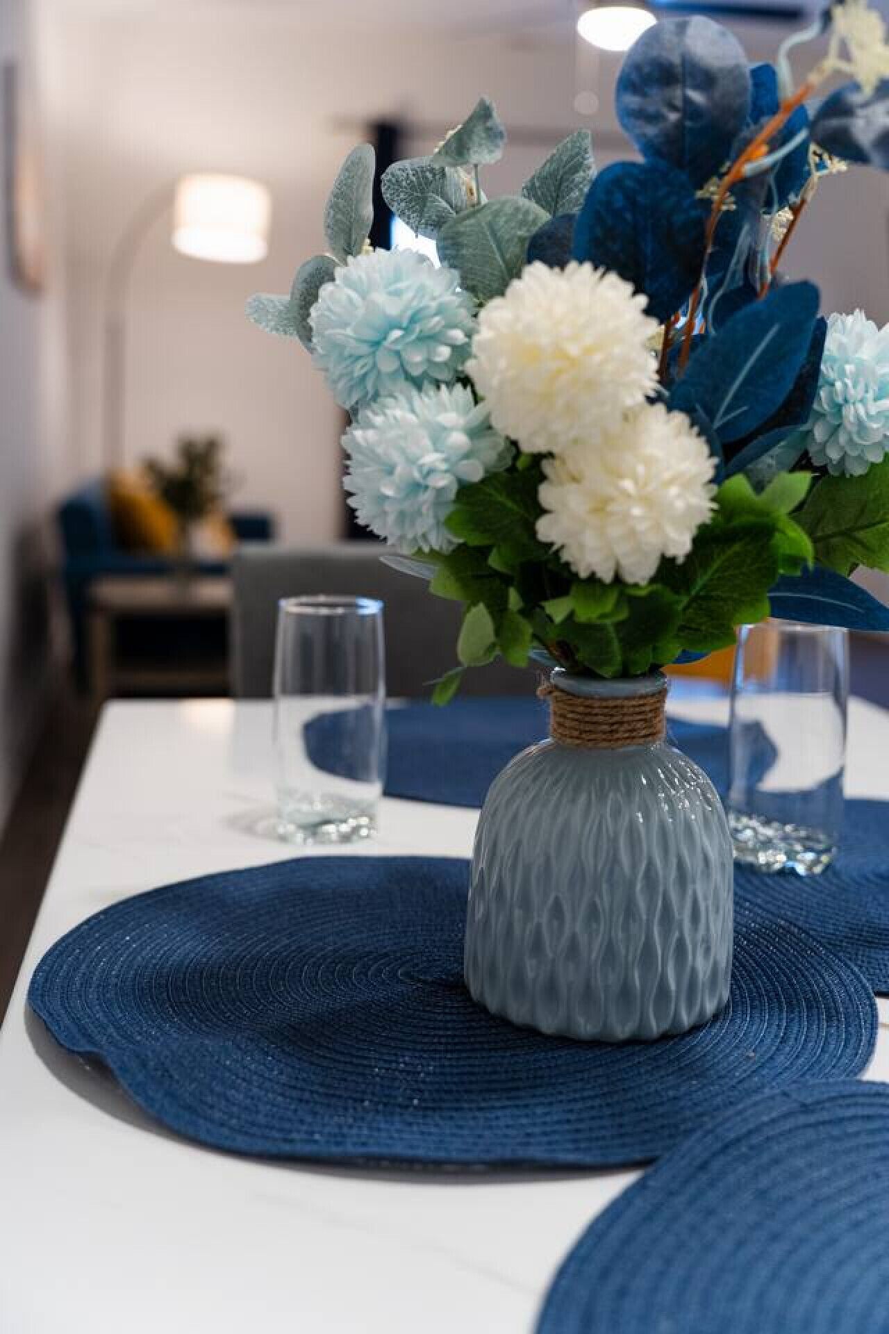 A dining table with a vase of flowers, blue placemats, and clear glasses.