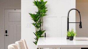 Una cocina con cubierta blanca, grifo negro y una planta en maceta. Hay tres taburetes de bar con patas negras y asientos blancos.