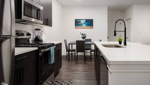 Una cocina moderna con armarios de madera oscura, electrodomésticos de acero inoxidable y una encimera blanca. Hay un área de comedor con una mesa y sillas, y un cuadro en la pared.
