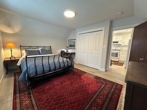 Ein Schlafzimmer mit Bett, Nachttisch, Schreibtisch, Stuhl, einem roten Teppich mit Muster und einem weißen Kleiderschrank. Durch eine offene Tür ist eine Küche zu sehen.