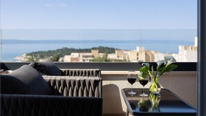 A balcony with a view of the sea and a cityscape. There is a sofa, a small table with two glasses of red wine, and a vase with white flowers.