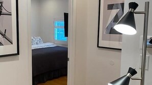 Um corredor estreito com piso de madeira que leva a um quarto com cama e janela. Há duas luminárias de chão pretas em cada lado do corredor.