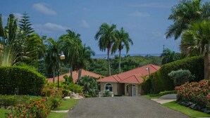 A residential area with a paved driveway leading to a house with a red-tiled roof, surrounded by lush greenery and palm trees. - Moradia com piscina privada em residência fechada, totalmente equipada (Sosúa)