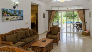 A spacious living room with a high ceiling, a painting on the wall, a wooden coffee table, and a view of the outdoor patio area. - Moradia com piscina privada em residência fechada, totalmente equipada (Sosúa)