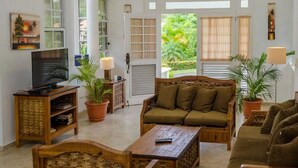 A spacious living room with a high ceiling, large windows, and a view of greenery outside. There are multiple seating areas with wooden furniture and a television on a stand. - Moradia com piscina privada em residência fechada, totalmente equipada (Sosúa)