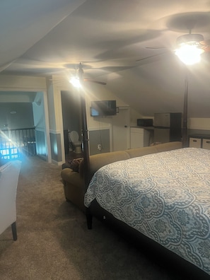 A bedroom with a large bed, a ceiling fan, a TV, and a kitchenette.
