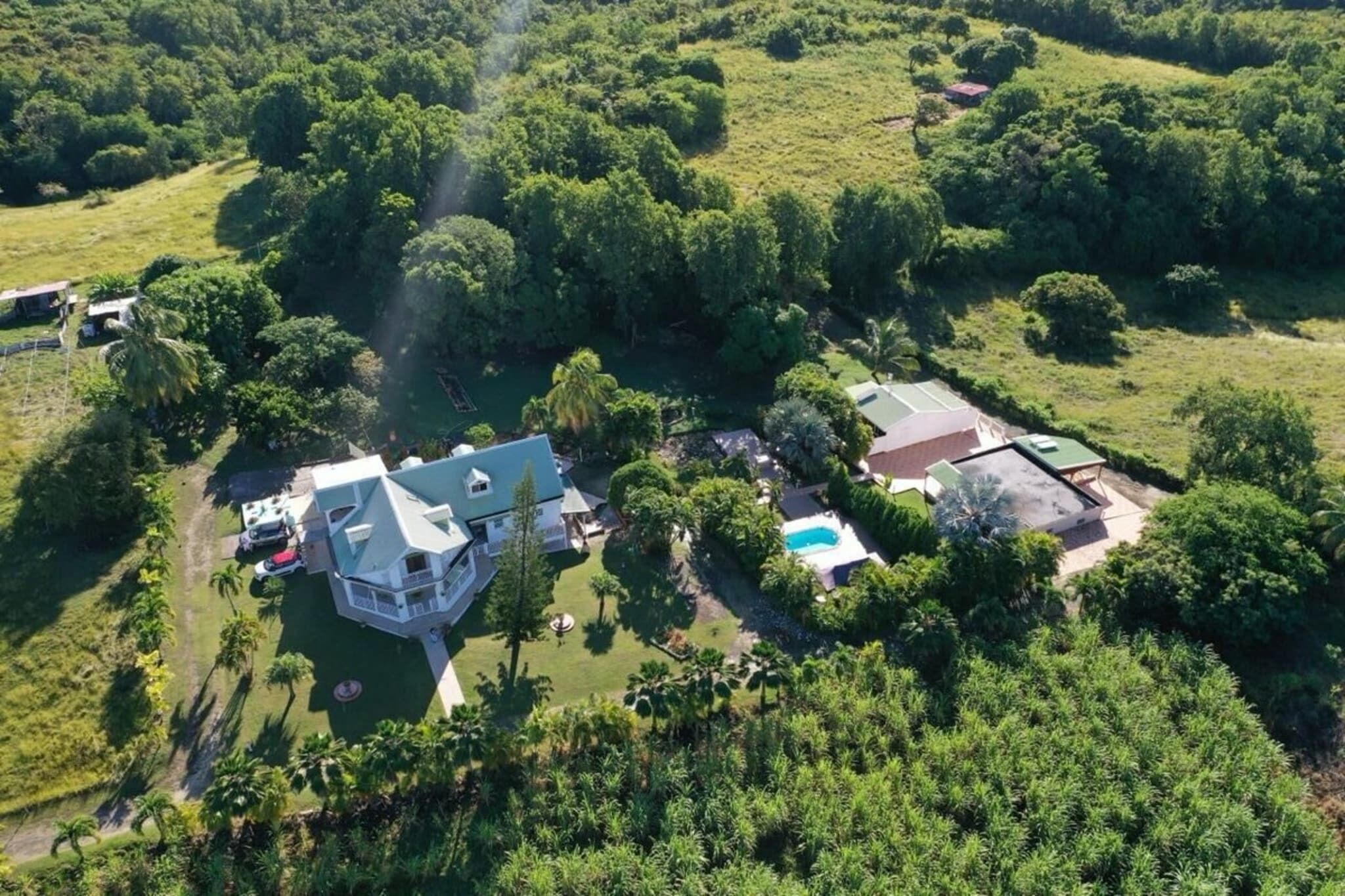 An aerial view of a property with a large house, a swimming pool, and a tennis court surrounded by lush greenery.