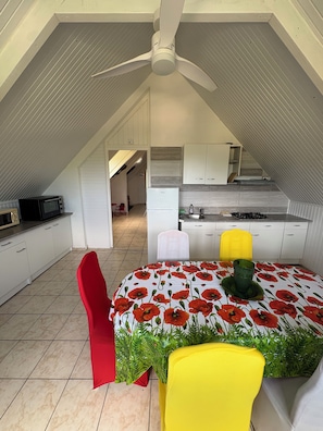 Un comedor con una mesa cubierta por un mantel rojo y blanco con motivos florales, dos sillas, un ventilador de techo y una cocina al fondo.