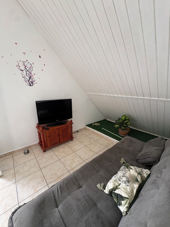 Una habitación con un sofá gris, un televisor sobre un mueble de madera, una planta pequeña en maceta y un adhesivo de árbol en la pared.