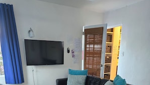 Un salon avec un canapé en cuir noir, une table basse en bois, un téléviseur fixé au mur et une fenêtre avec des stores.