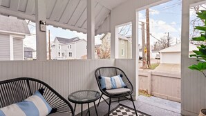 Eine überdachte Veranda mit zwei schwarzen Korbstühlen, einer mit einer Streifenauflage und der andere mit einer blau-weißen Auflage, sowie einem kleinen runden Tisch.