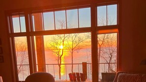 A room with a large window overlooking a sunset, two armchairs, a small table, and a lamp.
