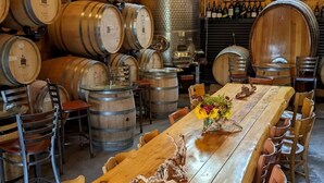 A rustic interior with wooden barrels, a long wooden table with chairs, and a bar area with bottles.