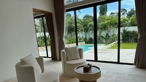 Amplia habitación con dos sillones beiges y una mesa de centro redonda, ventanas grandes que ofrecen vistas a una piscina y abundante vegetación.