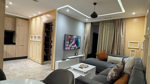 A modern living room with a grey sofa, a white coffee table, and a TV mounted on the wall. - Luxury apartment in Agdal free Netflix and garage (Rabat)