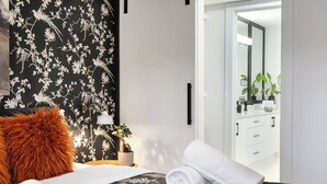 Une chambre à coucher avec un papier peint à motifs floraux noir et blanc, une table de chevet blanche ornée d’une plante, une serviette blanche roulée et une couvre-lit noire matelassée.