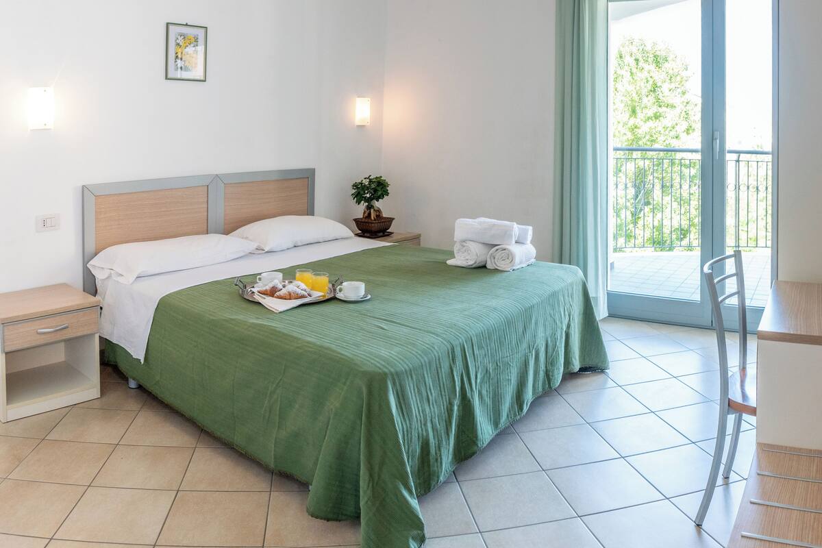 Quarto de hotel com cama, mesa de cabeceira, cadeira, mesa, varanda com vista e planta em vaso.