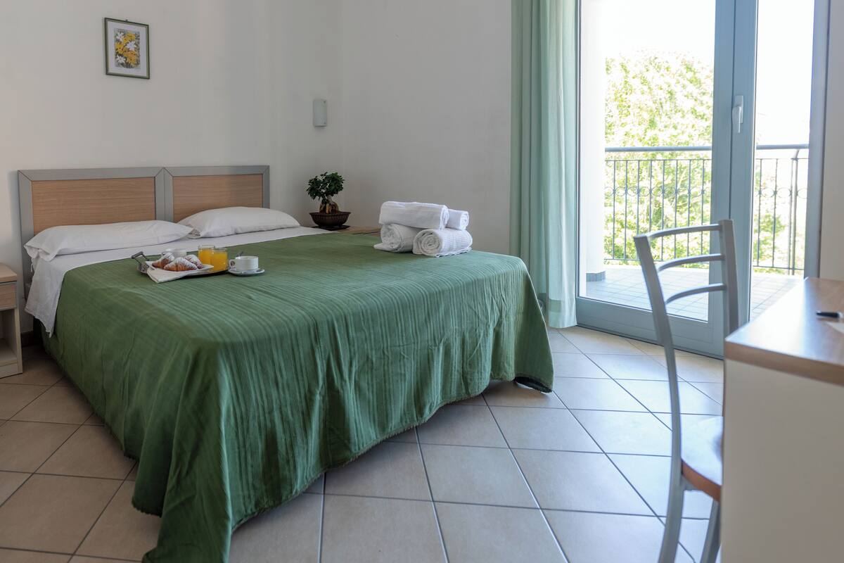 Quarto de hotel com uma cama, uma pequena mesa com cadeira, um vaso de planta e uma varanda com guarda-corpo.