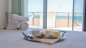A neatly made bed with a tray of croissants and orange juice, a rolled towel, and a cup, with a balcony view of the beach and ocean.