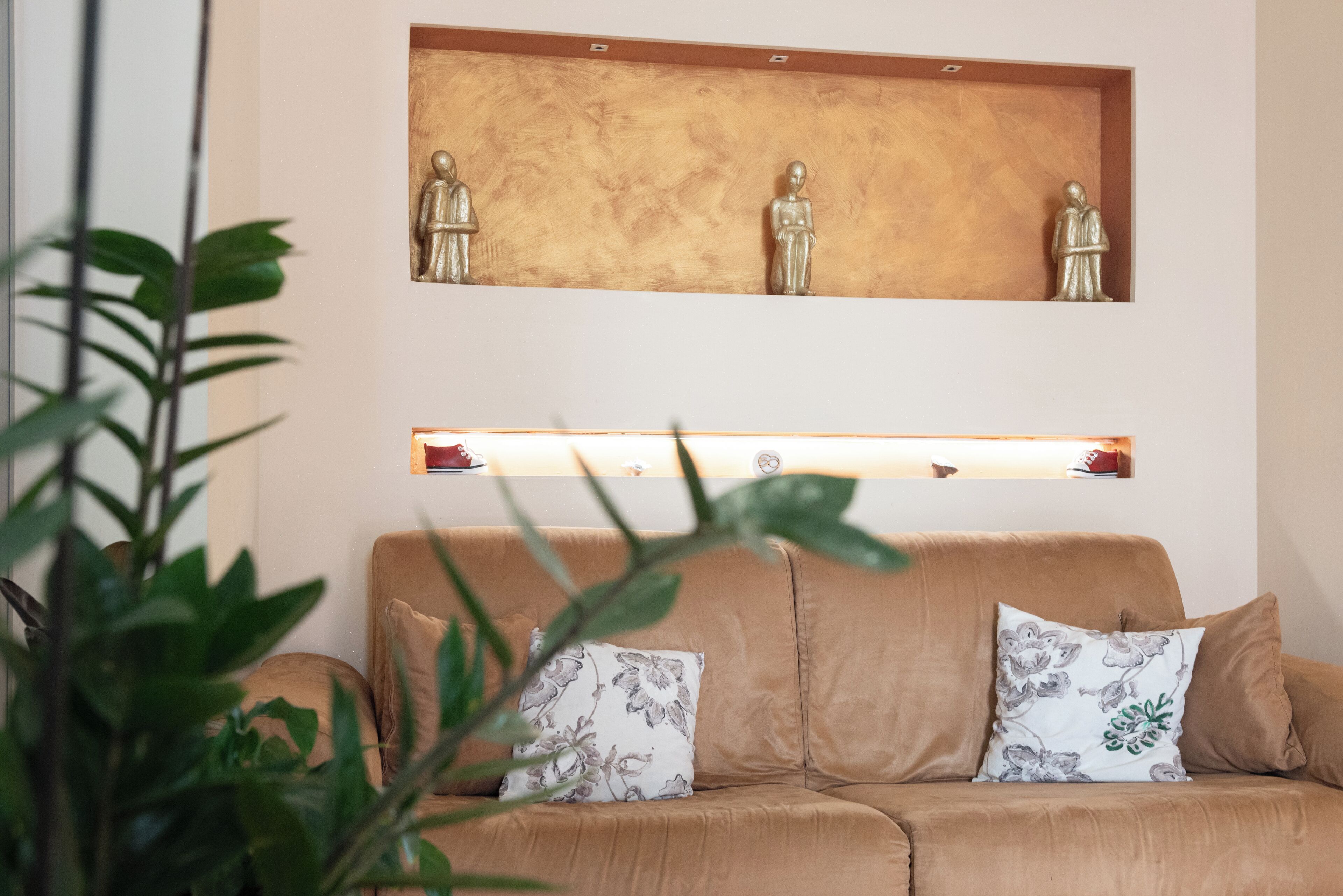 Un sofá color beige con cojines florales, un estante de madera con tres estatuas y una planta en primer plano.