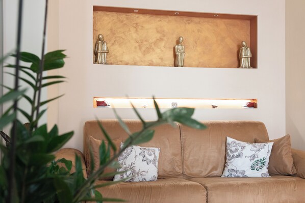 A beige sofa with floral cushions, a wooden shelf with three statues, and a plant in the foreground. - Altamira Residence (Roseto degli Abruzzi)