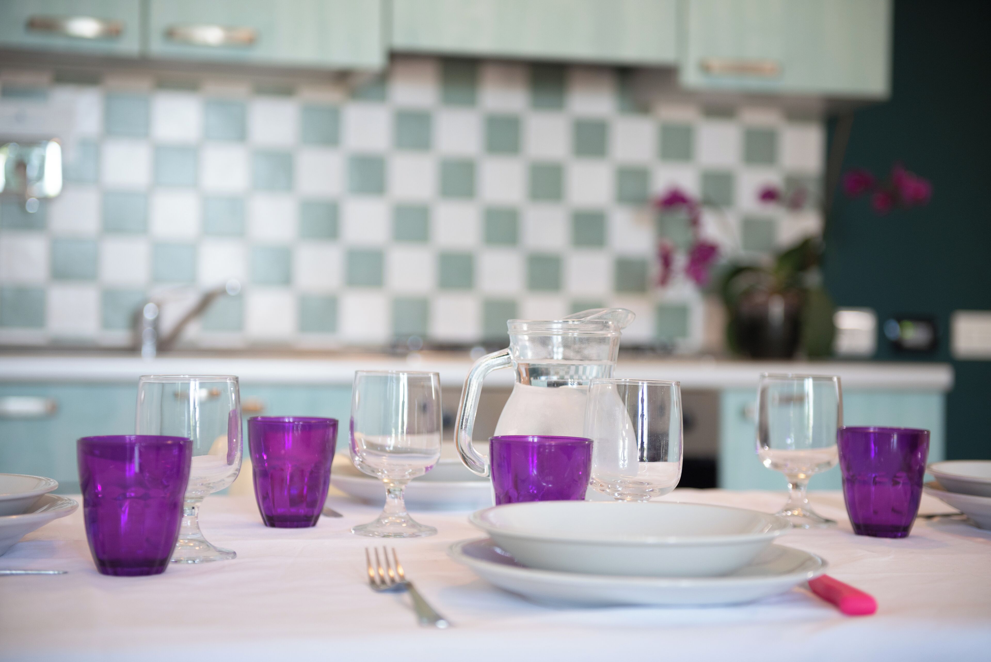 Un tavolo apparecchiato con bicchieri viola, un boccale e piatti in una cucina con mobili azzurro mare e uno schienale a scacchi.