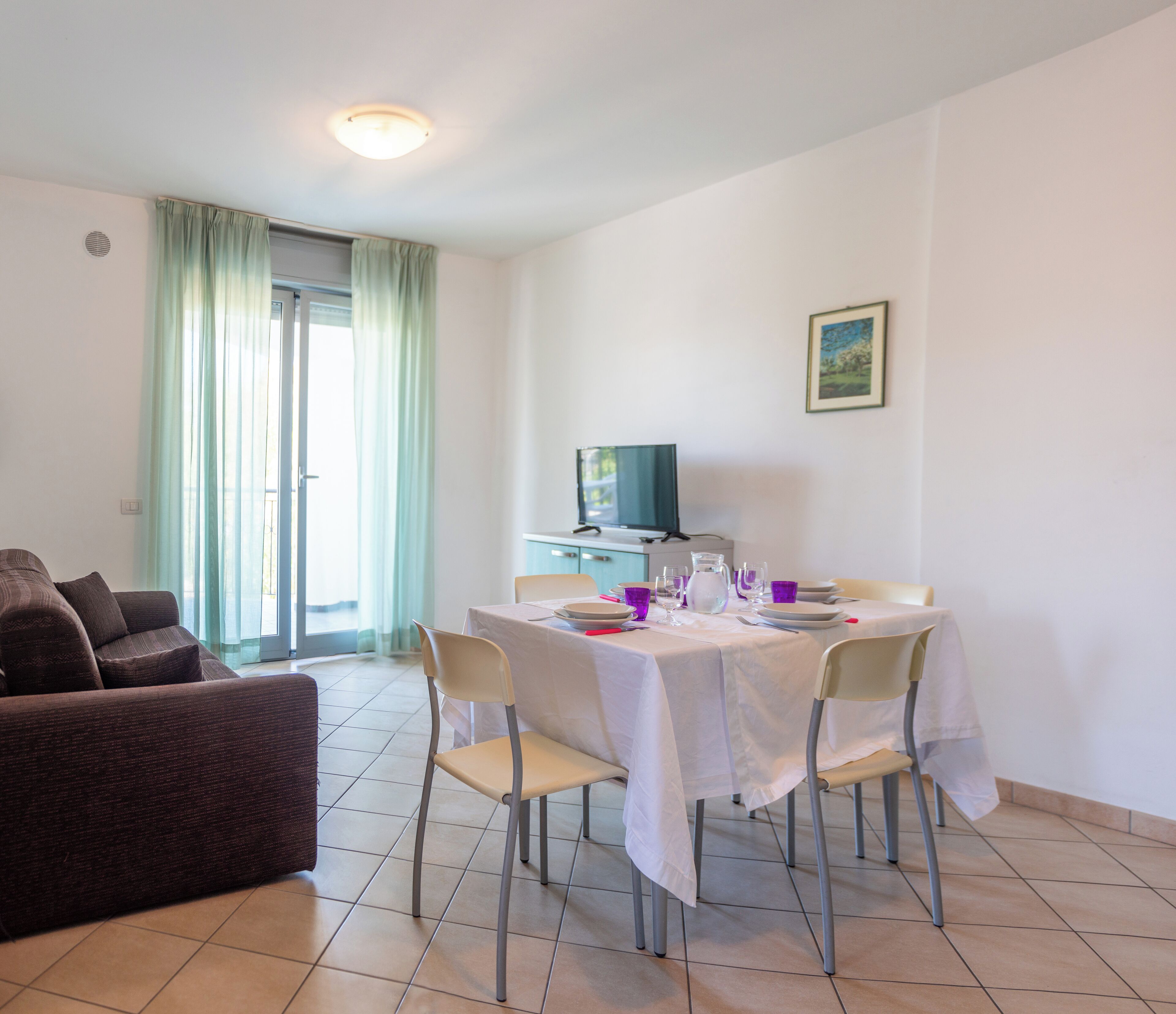 A dining area with a table set for four, a sofa, a TV, and a window with curtains.
