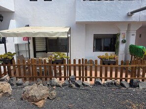 A building with a white exterior, a wooden fence, potted plants, and a tiled patio area. - Casa Samba, 2 Bedrooms and Pool (La Oliva)