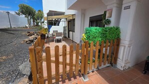A white building with a wooden fence, a patio with a table and chairs, and a green bush. - Casa Samba, 2 Bedrooms and Pool (La Oliva)