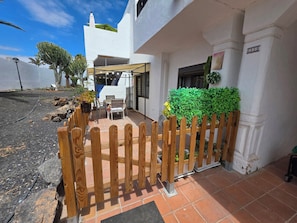 A white building with a wooden fence, a patio with a table and chairs, and a green bush. - Casa Samba, 2 Bedrooms and Pool (La Oliva)