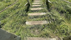 Eine Holztreppe führt zu einem Strand mit freiem Blick auf das Meer und ferne Hügel.