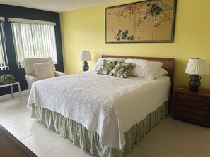 Een net opgemaakt bed met een witte deken en een groene sierrok, een nachtkastje van hout met een lamp, een stoel met een gestreept kussen, en een muur met een bloemschilderij.
