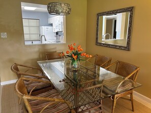 Een eetruimte met een glazen tafel, omringd door rieten stoelen, een vaas met oranje bloemen en een spiegel aan de muur.