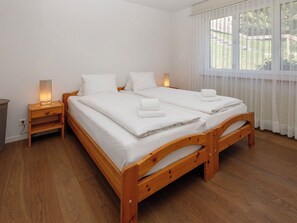 A double wooden bed with white linens and towels, a bedside table with a lamp, and a window with sheer curtains.