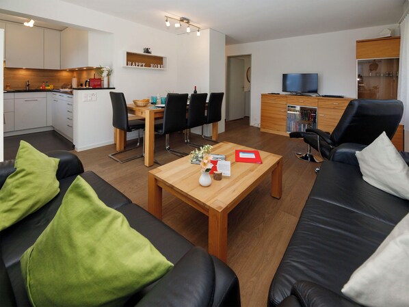 A modern living room with a sofa, a coffee table, and a TV. There is a dining area with chairs and a table, and a kitchen with cabinets and a countertop.