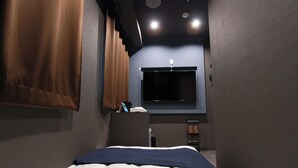 A bedroom with a bed, a window with curtains, a small table with a phone and a bottle, and a shoe rack. - Epoch inn Nakasu (Fukuoka)