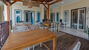 Un espacio interior amplio con mesas y sillas de madera, un techo alto con vigas de madera y grandes puertas de cristal que dan a otra habitación.